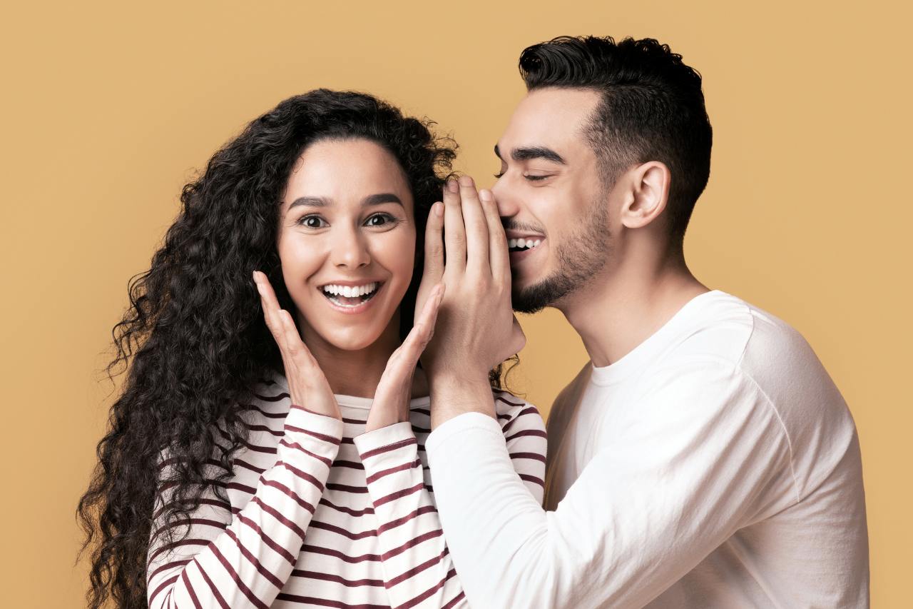 smiling man whispering to a pretty woman with an excited look on her face